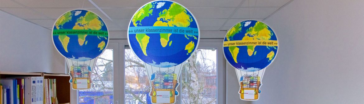 Three cardboard hot-air balloons bearing the words "Our classroom is the world" hang in front of a window.